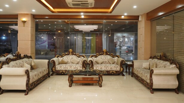 A spacious lobby with ornate sofas and a central coffee table, a chandelier, and a view of a dining area through a glass partition. - The gorbandh palace (Ankleshwar)