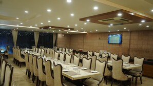A spacious dining area with multiple tables and chairs, a television mounted on the wall, and a tiled floor. - The gorbandh palace (Ankleshwar)