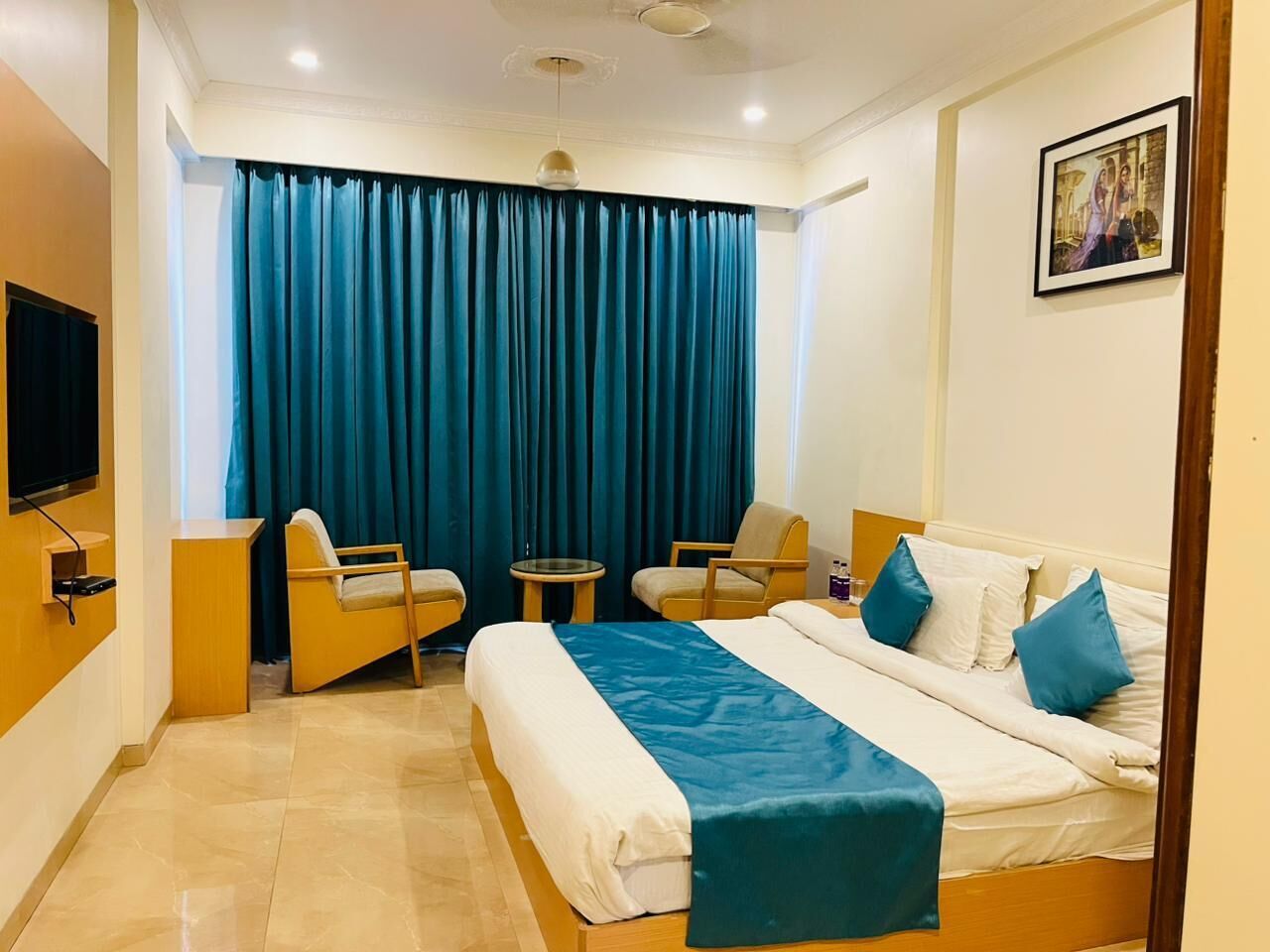 A hotel room with a bed, two armchairs, a small table, a TV, and a large window with curtains.