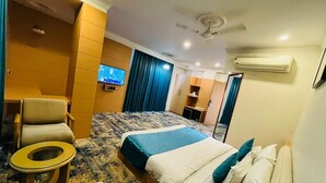 A hotel room with a bed, a chair, a small table, a TV, and a ceiling fan. - The gorbandh palace (Ankleshwar)