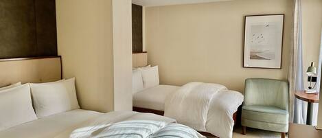 A hotel room with two beds, a chair, a small table with a lamp, and a framed picture on the wall.