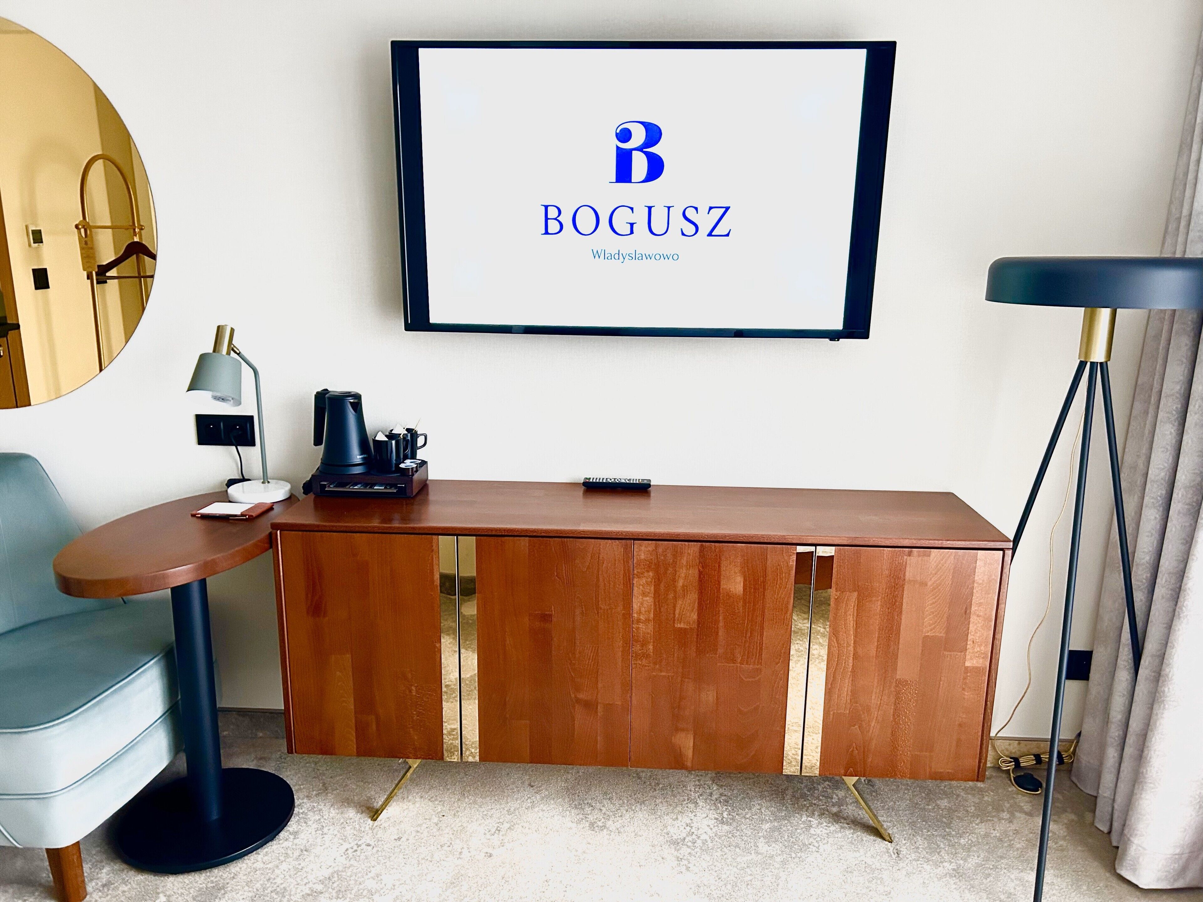 Una habitación de hotel moderna con una mesa consola de madera, un televisor de pantalla plana mostrando el logotipo de BOGUSZ, una lámpara de escritorio, una cafetera y una silla azul.