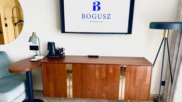 Una habitación de hotel moderna con una mesa consola de madera, un televisor de pantalla plana mostrando el logotipo de BOGUSZ, una lámpara de escritorio, una cafetera y una silla azul.