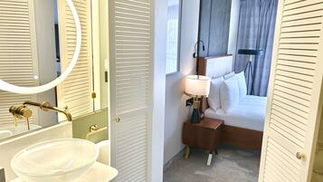 Habitación de hotel con baño que incluye un lavabo, espejo y tocador. La habitación también tiene una cama con mesita de noche, lámpara y cortinas.