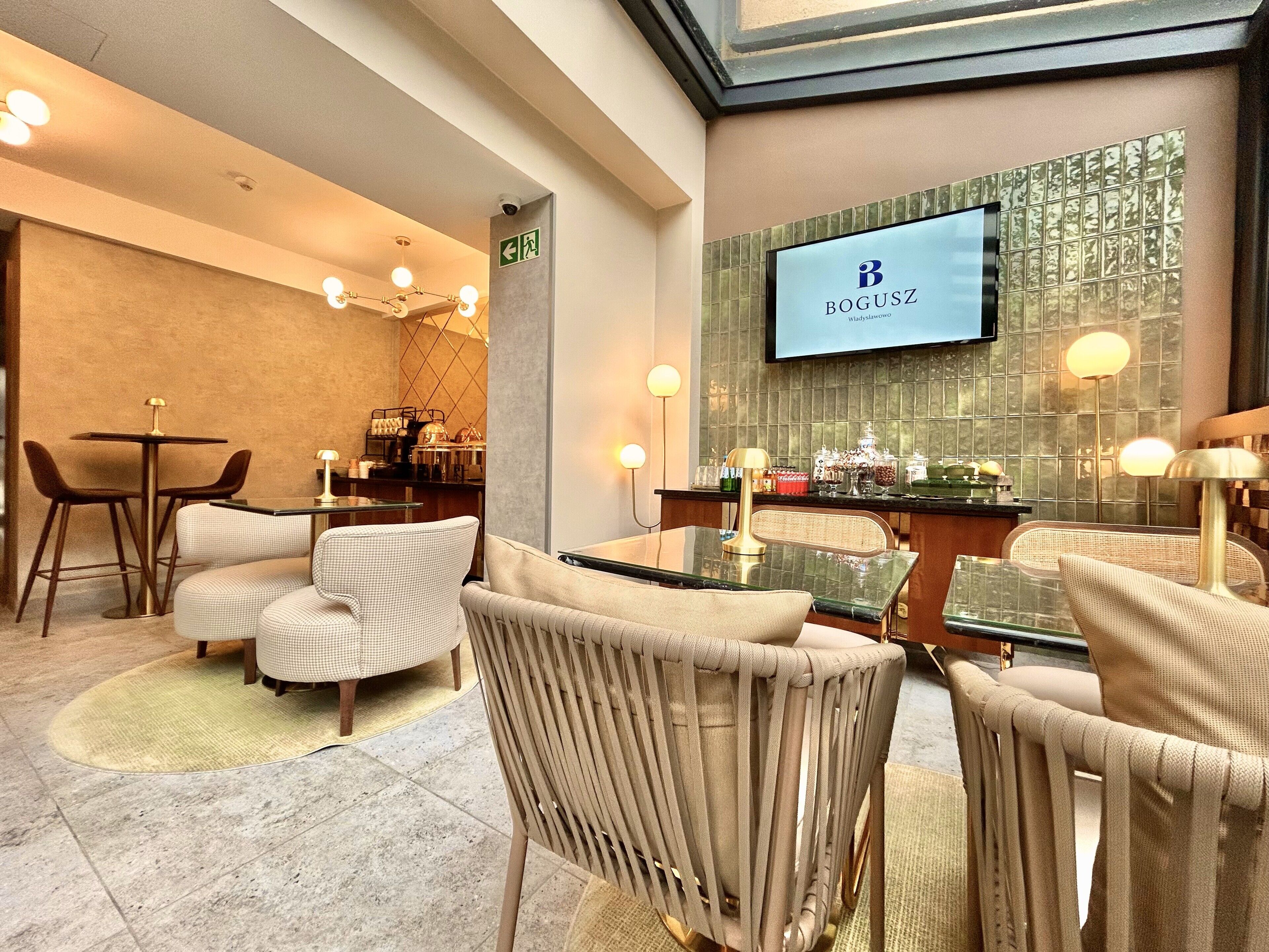 A modern hotel lobby with a reception desk, comfortable seating, a television screen displaying the hotel name, and a decorative wall with a geometric pattern.