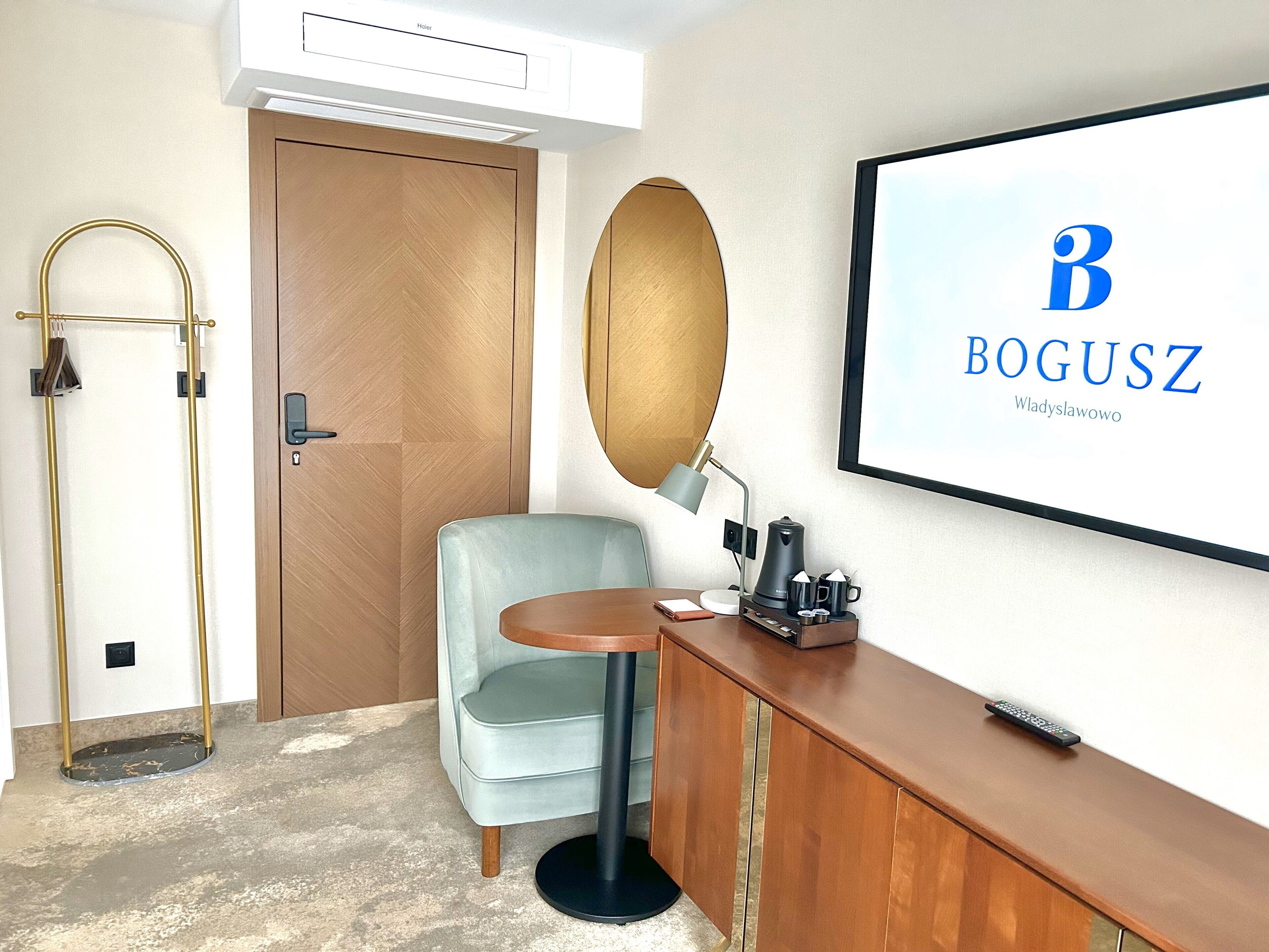 A hotel room with a wooden door, a light blue armchair, a round mirror, a coffee maker, and a framed 'BOGUSZ' sign on the wall.