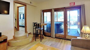 A room with a wooden coffee table, a sofa, a TV mounted on the wall, a dining table with chairs, and a view of the outside through a glass door. - Pinon Park A15 | Hot Tub, Great Location (Ruidoso)