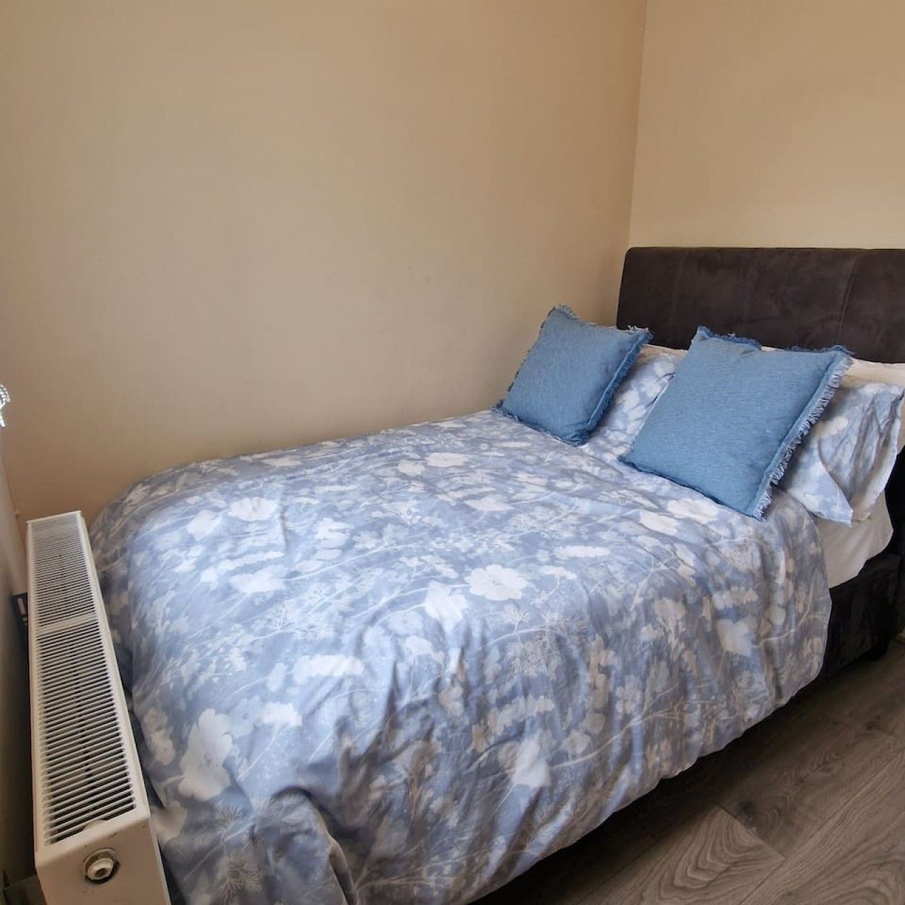 A single bed with a floral bedspread, a dark headboard, and a wall-mounted air vent.