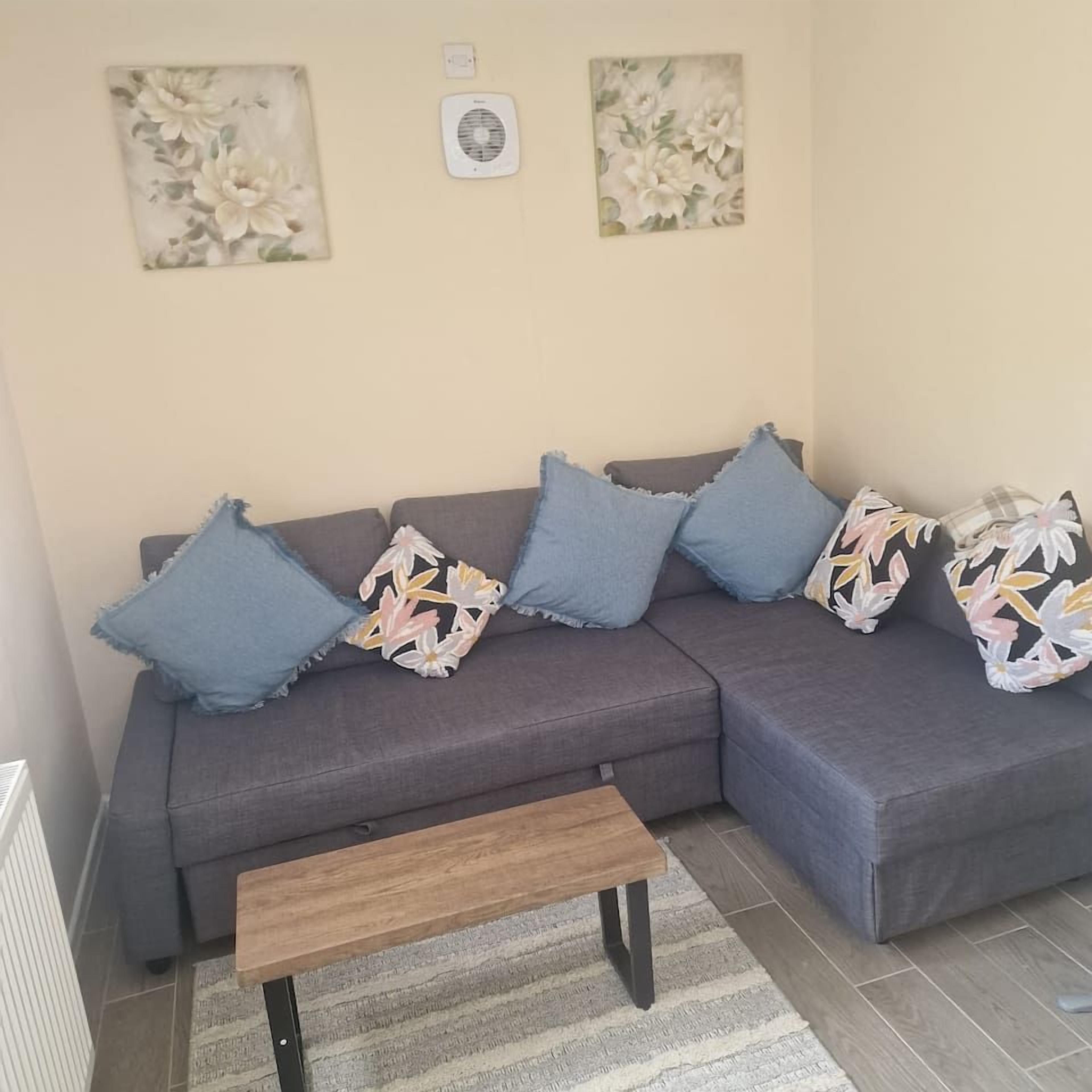 Een grijze bank met blauwe en bloemrijk bedekte kussens, een houten bankje en een gestreept vloerkleed.