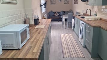 Een kleine keuken met een magnetron, spoelbak en wasmachine, die uitkomt in een zithoek met een bank en tafel.