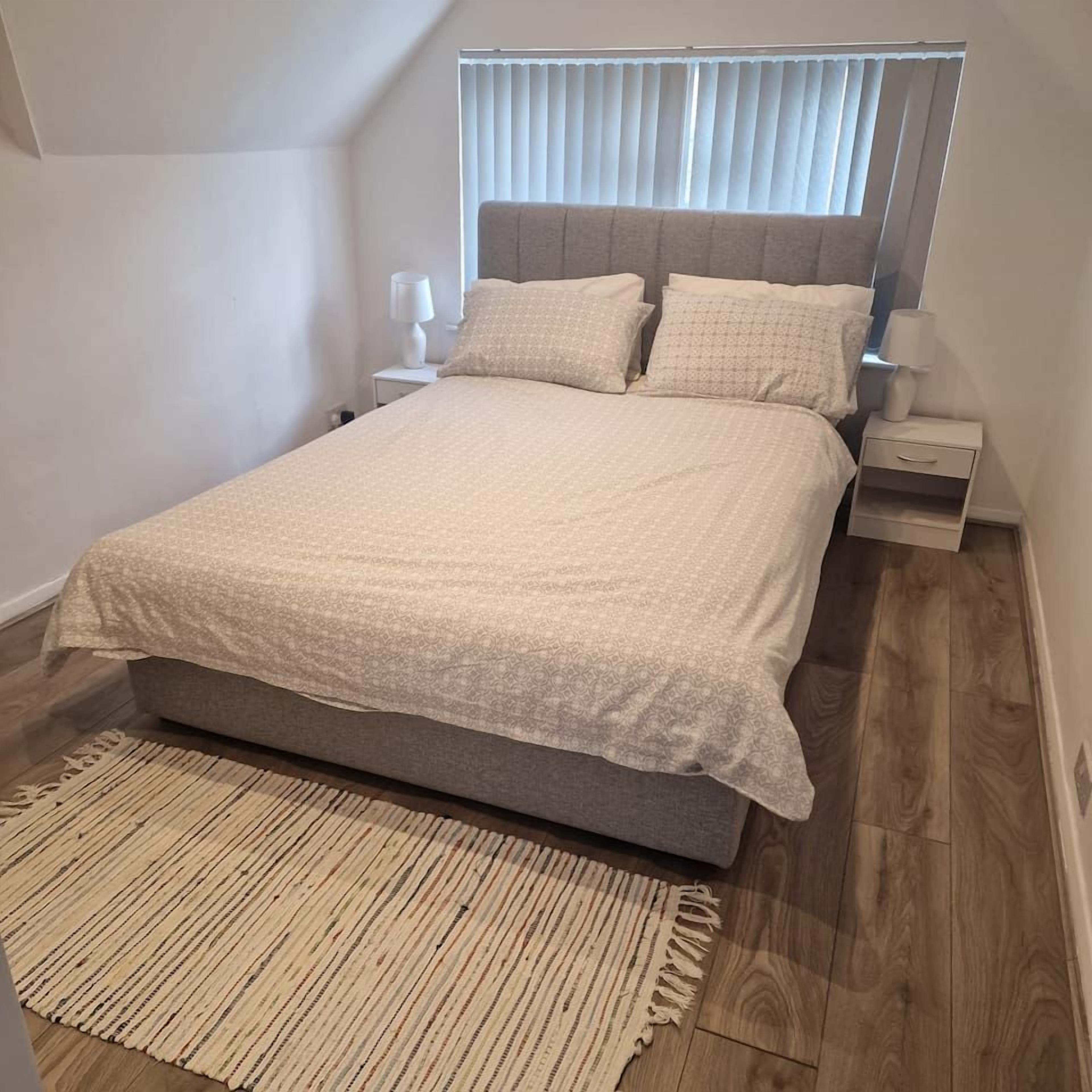 Een slaapkamer met een bed, nachtkastjes, een bedtafel, een raam met jaloezieën en een kamerbreed tapijt met dessin.