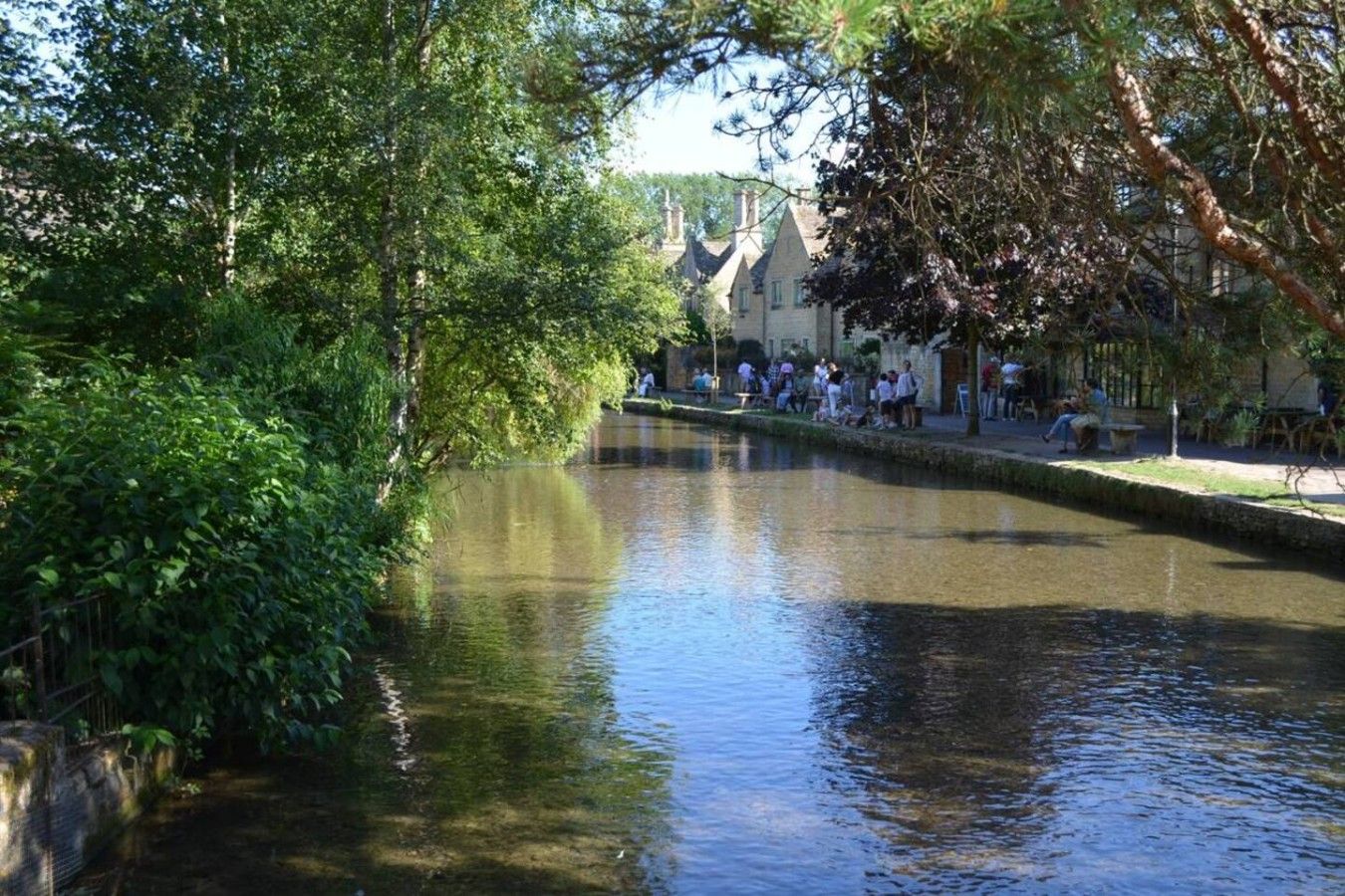 Une rivière paisible, avec un pont de pierre, des arbres et un bâtiment au bord duquel se trouvent des personnes assises.