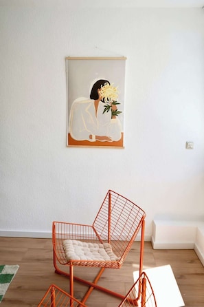Ein Zimmer mit weißer Wand, einem gerahmten Kunstwerk mit einer Person, die eine Blume hält, und einem einzigartigen orangefarbenen Drahtstuhl.