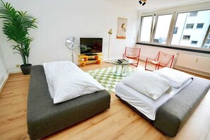 Ein Zimmer mit zwei Betten, einem Fernseher, einer Pflanze und einem Fenster mit Blick auf Gebäude.