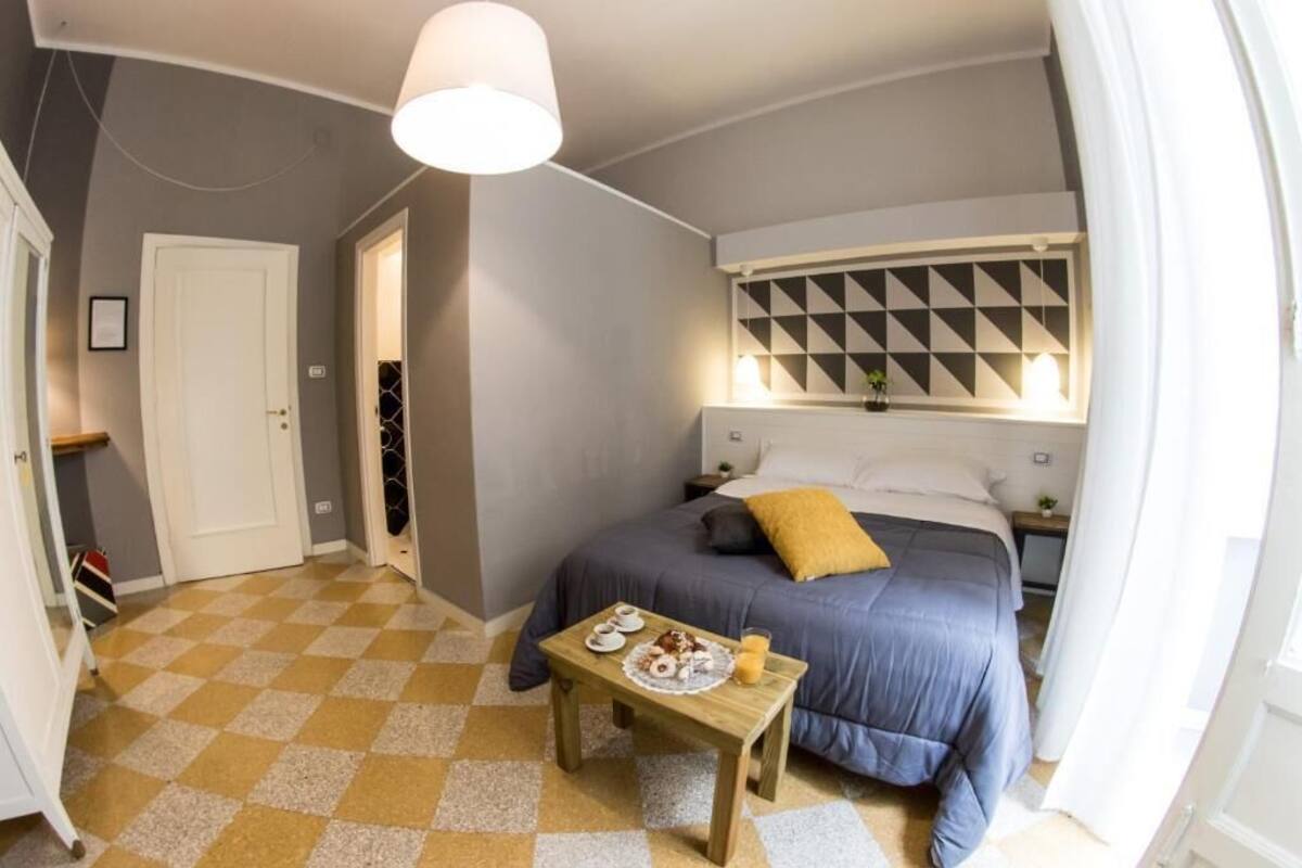 Quarto com chão xadrez, cama com colcha azul e travesseiro amarelo mostarda, mesa de cabeceira com prato de comida, e parede com padrão geométrico acima da cama.