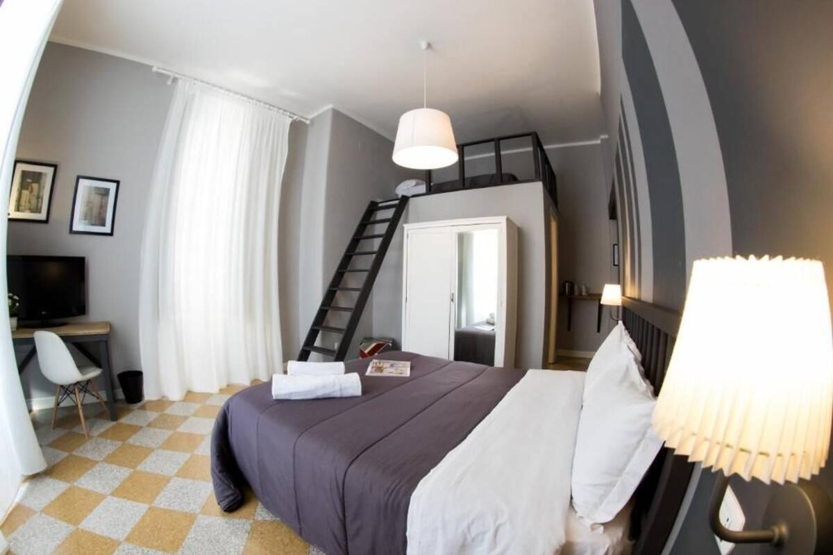 Quarto de hotel com cama, escrivaninha, cadeira, televisão, escada e luminária.
