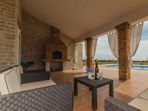 Une terrasse extérieure avec une cheminée en briques, un canapé, une table avec une bouteille et un verre, et une piscine en arrière-plan.