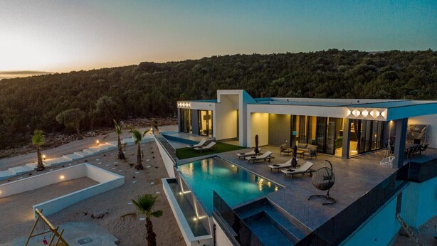 A modern villa with a swimming pool, outdoor seating, and a view of a forested hillside.