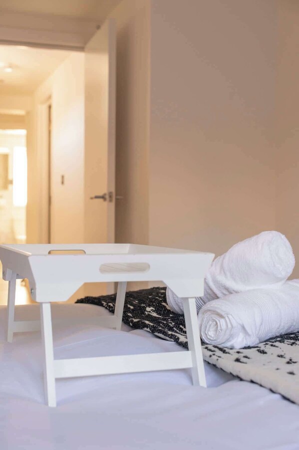 A hotel room with a bed, a white bedside table, and a rolled white towel.
