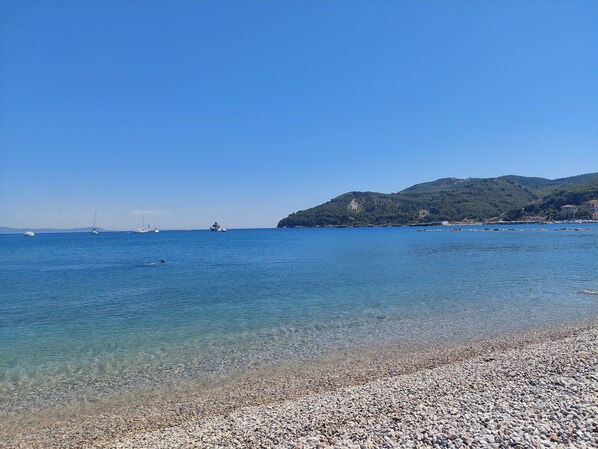 A pebbled beach with clear water and boats in the distance.