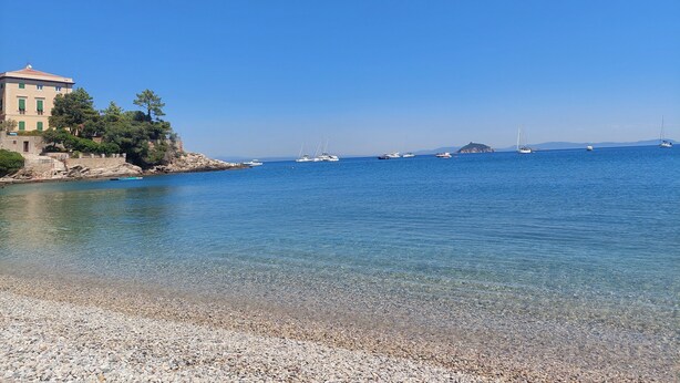 A coastal scene with a building on a rocky shore, clear blue water, and boats in the distance. - Apartment Cala Azzurra - with large garden and a stone's throw from the sea (Cavo)