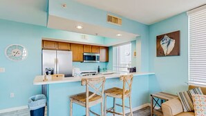 Cocina compacta con electrodomésticos modernos, zona de comedor con sillas de madera y un pequeño rincón de estar con sofá y cojines.