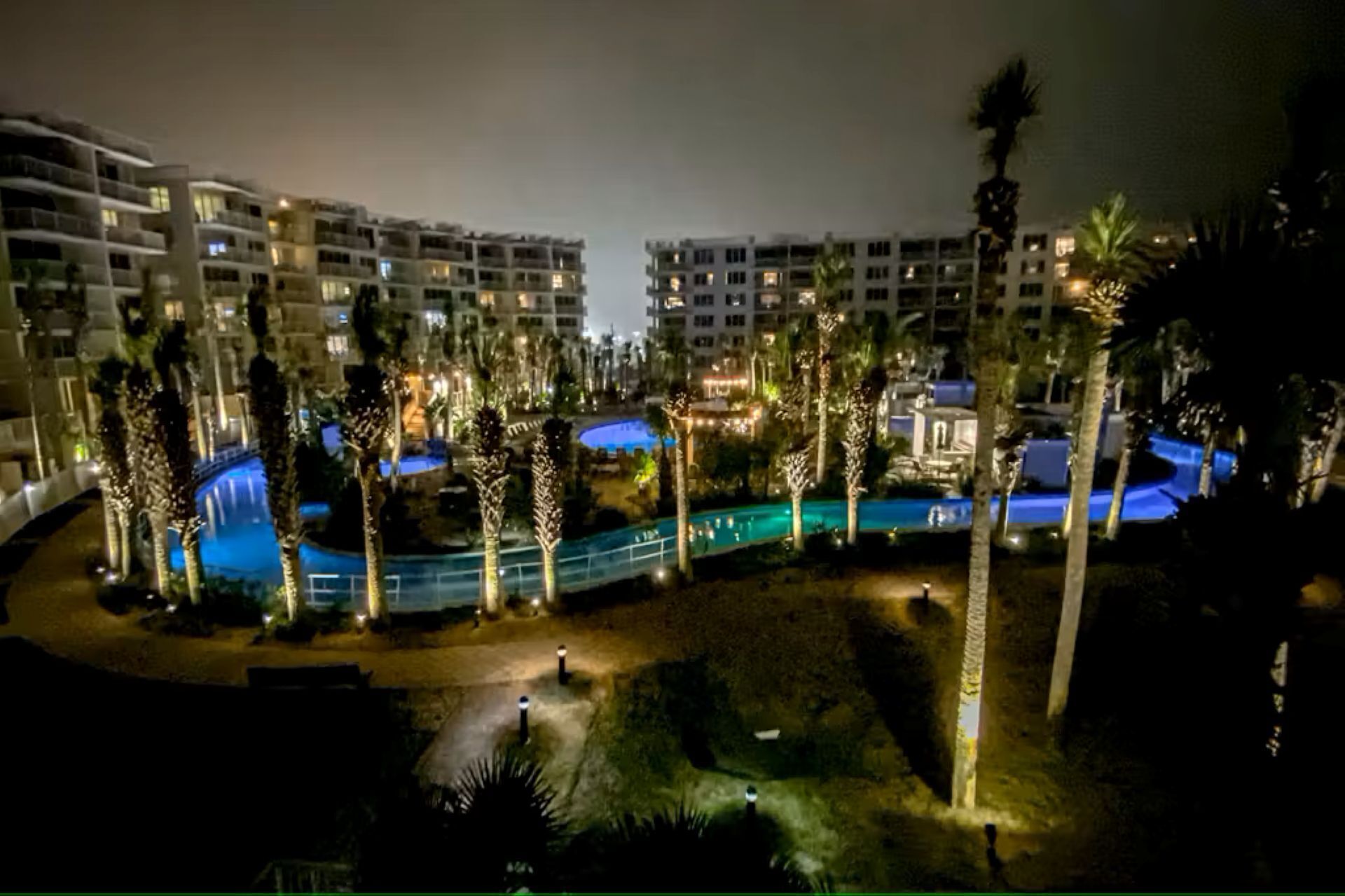 A multi-story hotel with a pool area surrounded by palm trees and illuminated by artificial lighting.