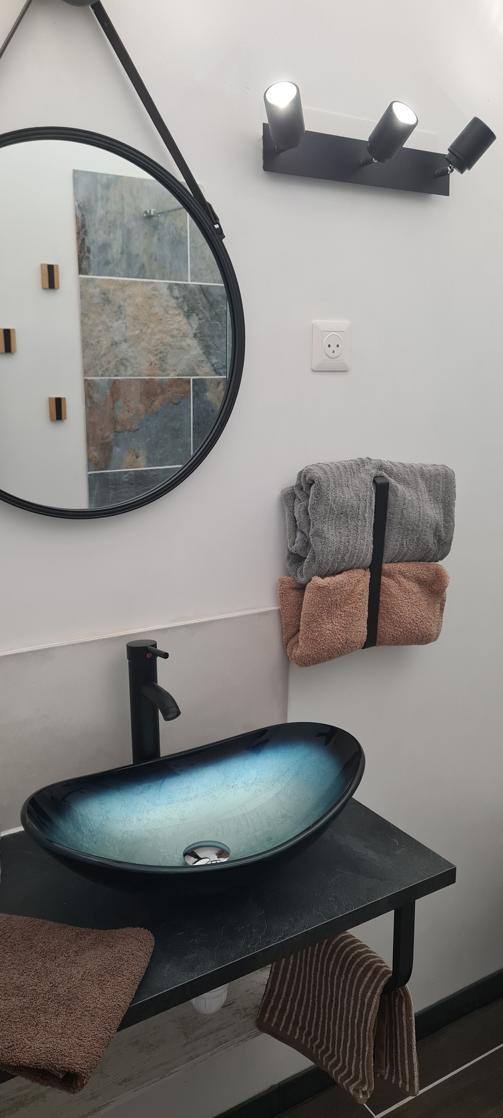 A bathroom with a round mirror, a black sink with a blue glass basin, and two towels hanging on hooks.
