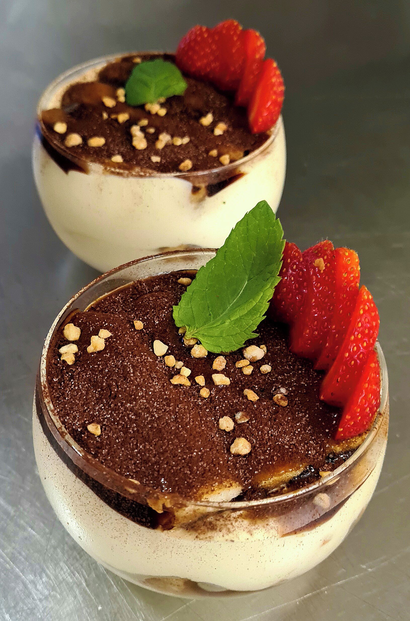 Two dessert cups with chocolate mousse, strawberries, and a mint leaf on a metallic surface.