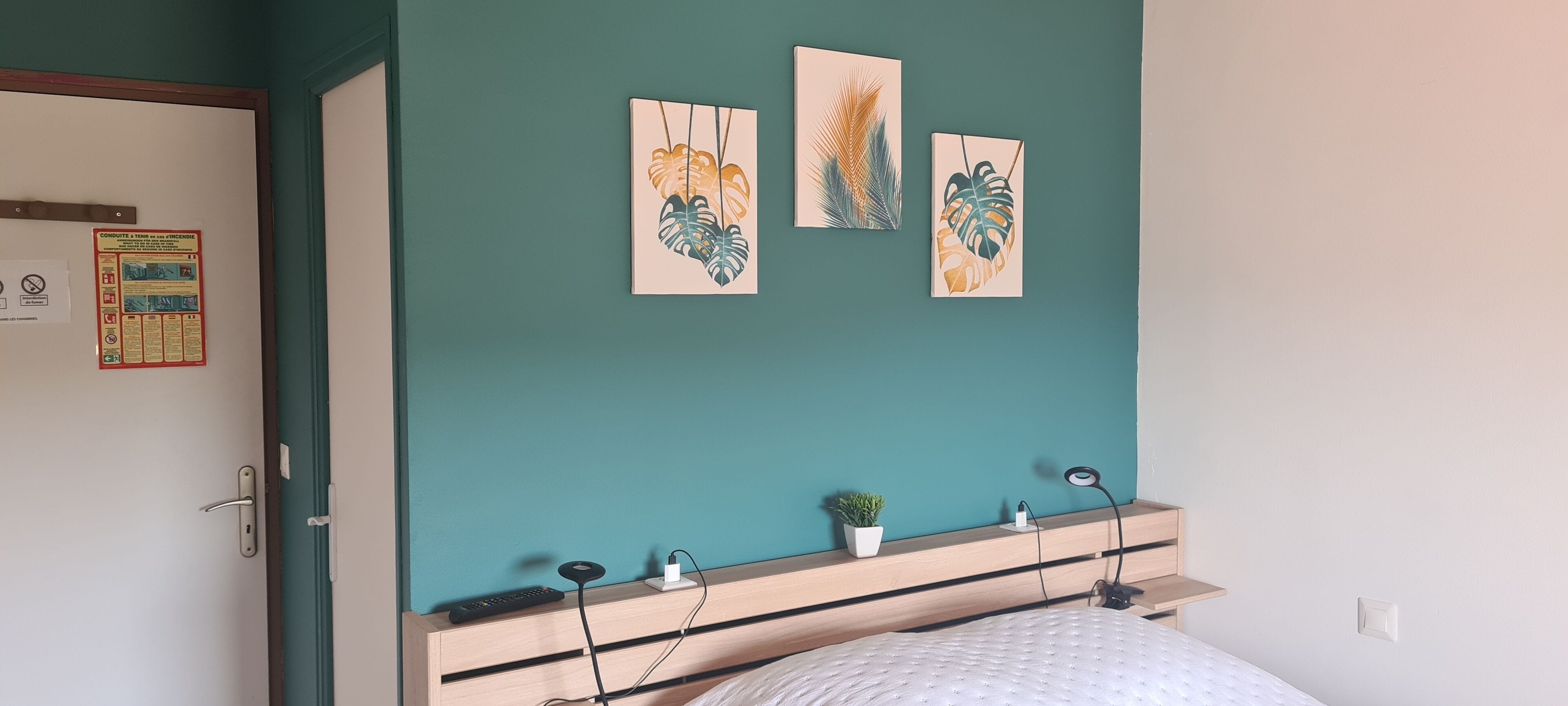Une chambre avec des murs de couleur turquoise, un lit recouvert d’une couverture blanche, une tête de lit en bois et trois œuvres d’art accrochées au mur, représentant des feuilles tropicales.