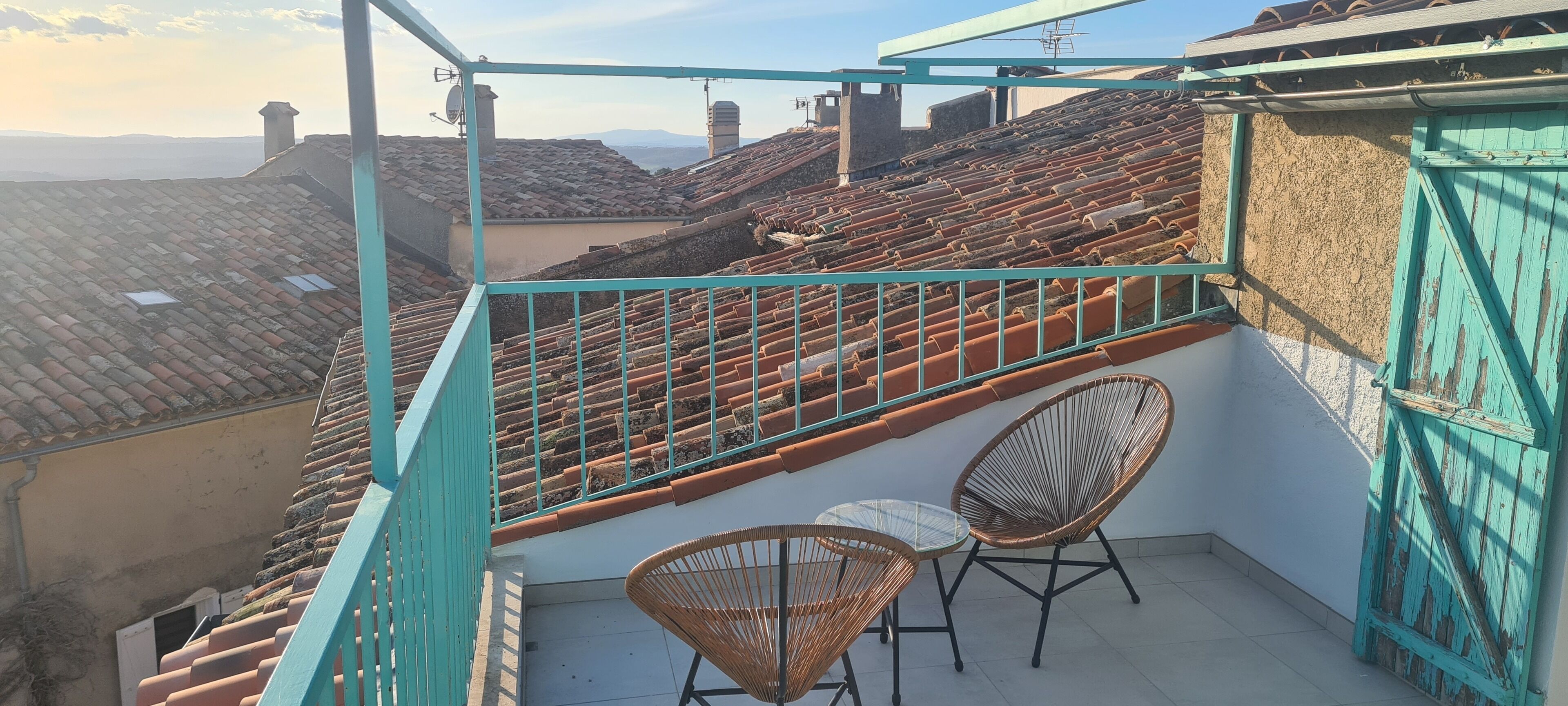 A balcony with a view of terracotta rooftops, a turquoise railing, and wicker chairs.