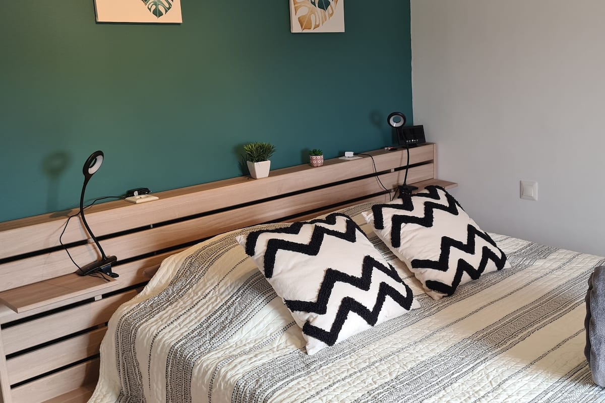 Ein ordentlich eingerichtetes Schlafzimmer mit einem Bett, zwei Kissen, einem Nachttisch, einer Lampe und einer kleinen Pflanze.