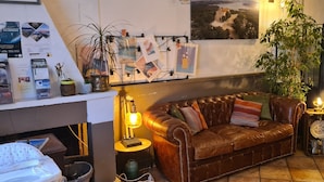 Een gezellige zithoek met een leren bank, een klein bijzettafeltje met een lamp, en een wand versierd met diverse objecten, waaronder een poster en een plant.