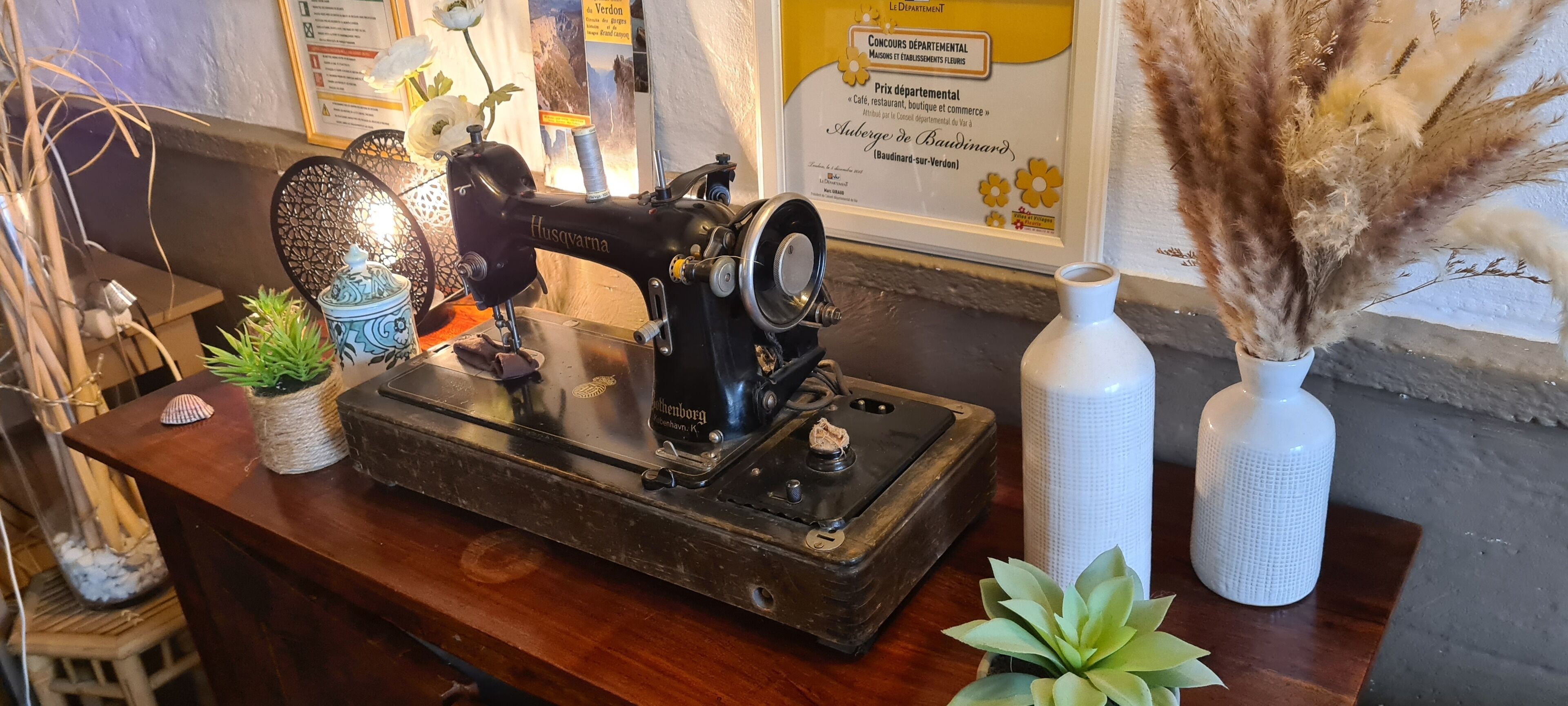 Una máquina de coser vintage Husqvarna sobre una mesa de madera, rodeada de objetos decorativos, incluyendo un jarrón de vidrio con plumas de pampa, una pequeña planta en maceta y una maceta de cerámica.