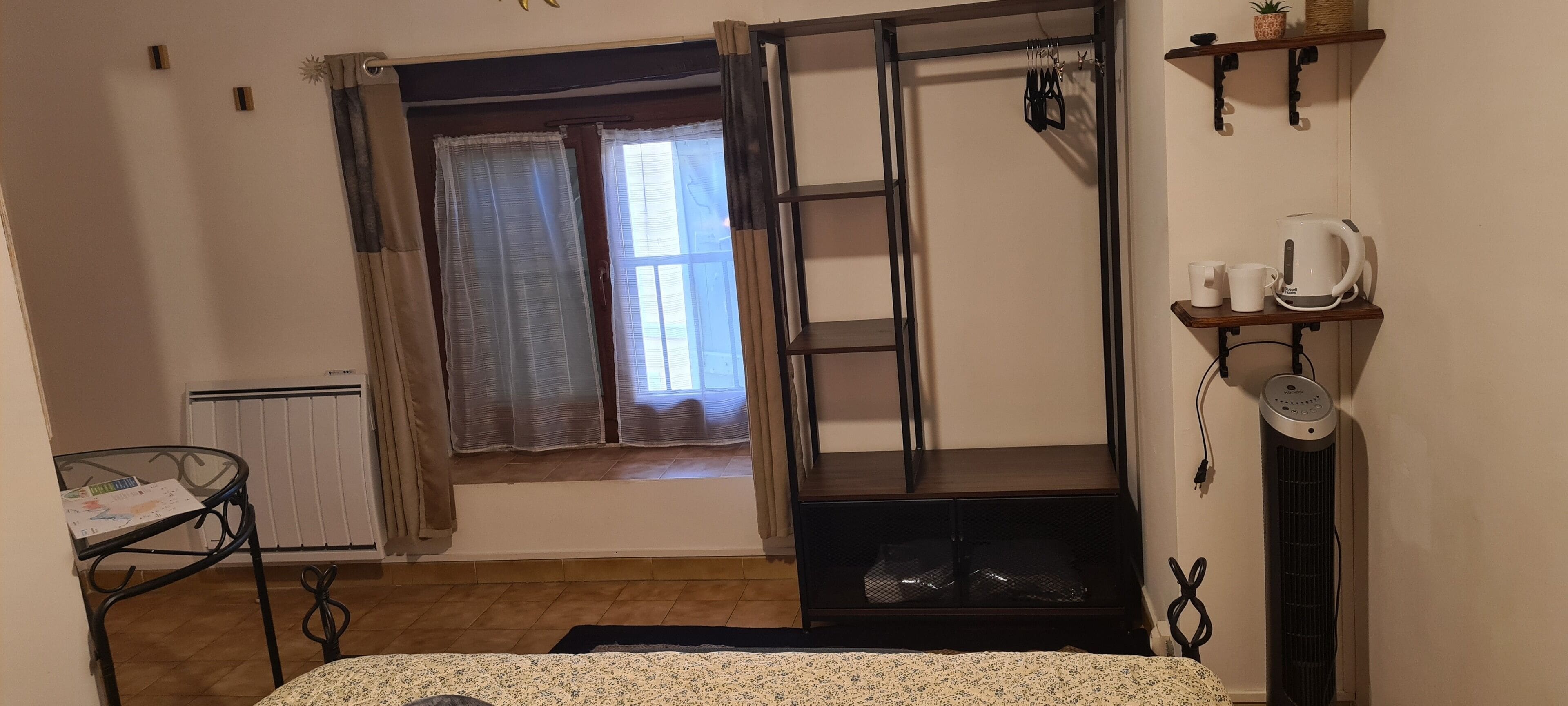 Una habitación con una cama, una ventana con cortinas, un radiador, una estantería con objetos y una mesa con un libro.