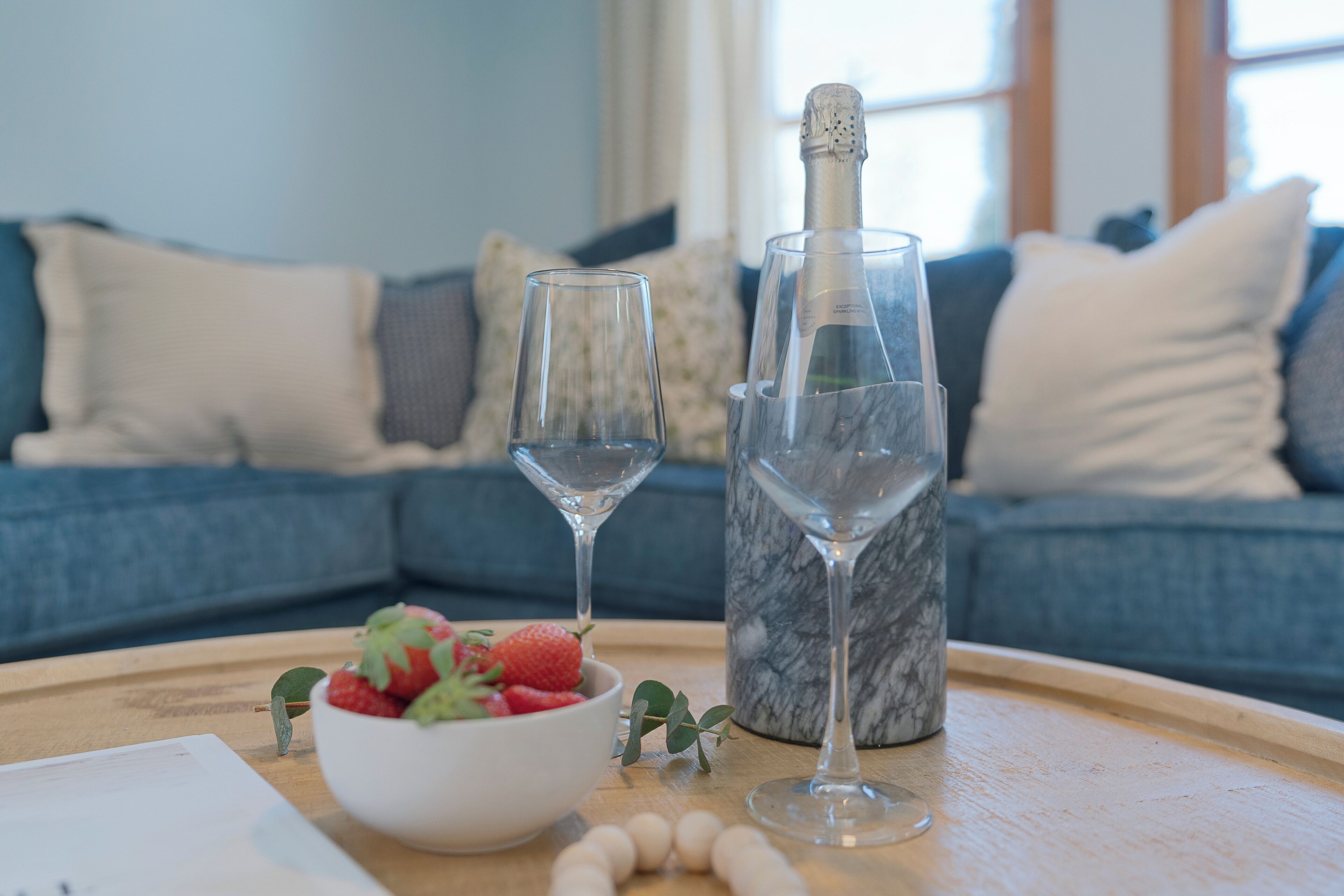 A tray with a bowl of strawberries, two wine glasses, and a bottle of champagne on a wooden table in a living room with a blue sofa in the background.