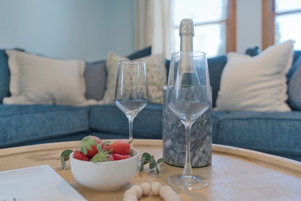 Una bandeja con un bol de fresas, dos copas de vino y una botella de champán sobre una mesa de madera en una sala de estar con un sofá azul al fondo.