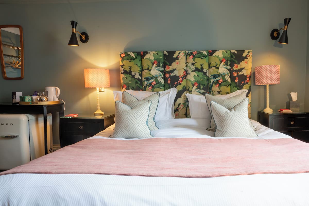 A neatly made bed with a floral headboard, two bedside lamps, and a small table with a kettle and cups.