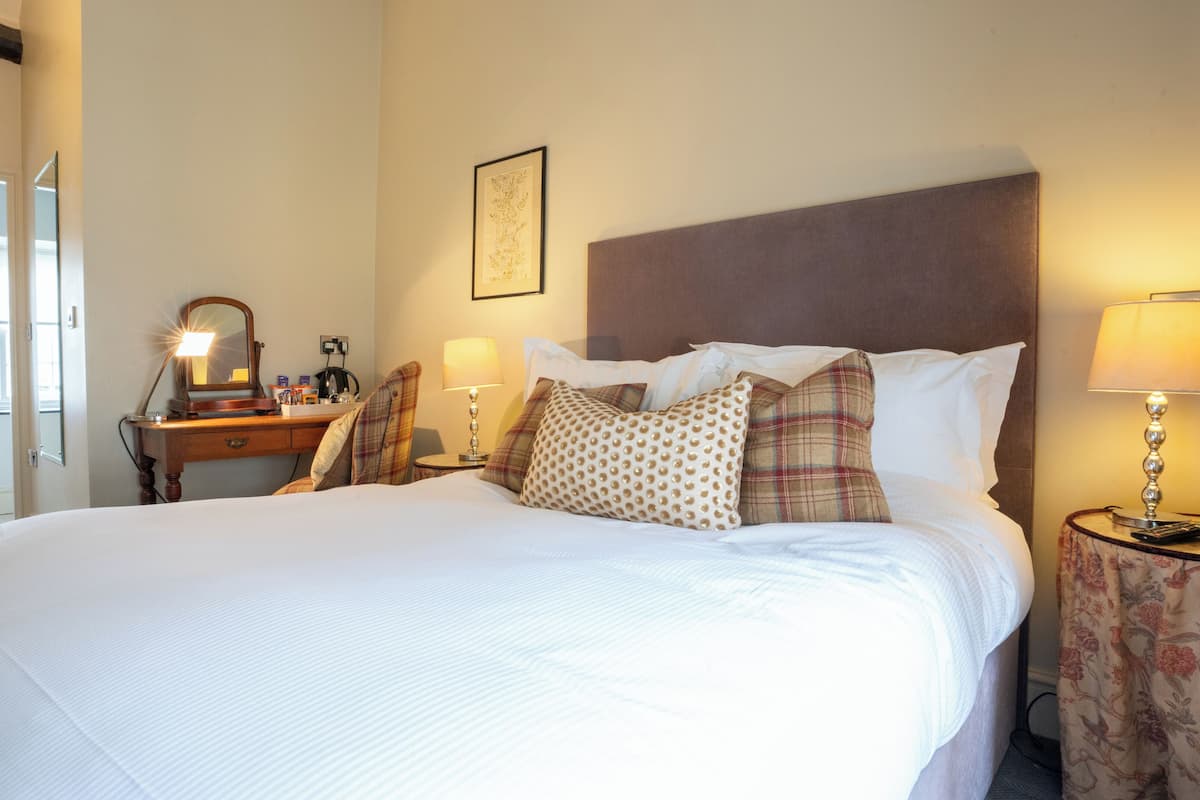 A neatly made bed with a headboard, a bedside table with a lamp, a dresser with a mirror, and a framed picture on the wall.