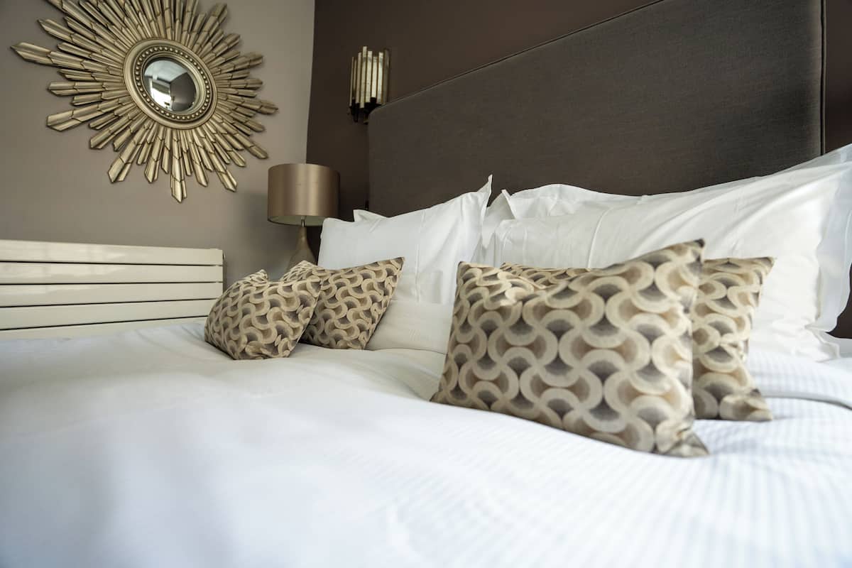 A neatly made bed with white linens and patterned pillows, a sunburst mirror on the wall, and a modern lamp.