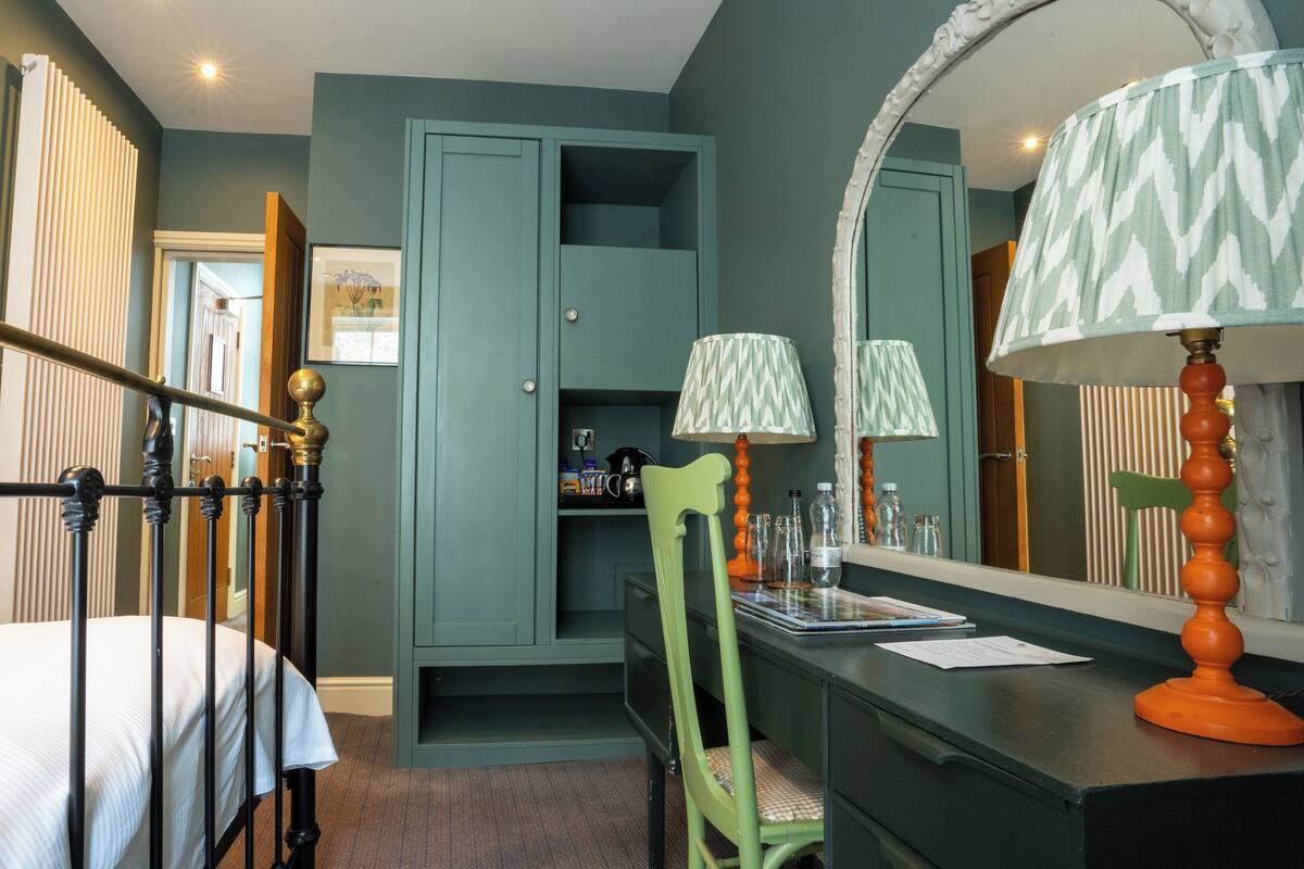 A bedroom with a four-poster bed, a desk with a chair, a wardrobe, and a large mirror.
