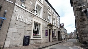 A stone building with a sign reading "Plato's" and a signboard for "Plato's" detailing opening hours and services. - Plato's (Kirkby Lonsdale)