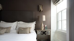 A neatly made bed with white linens and patterned pillows, a bedside table with a lamp, and a window with a view. - Plato's (Kirkby Lonsdale)