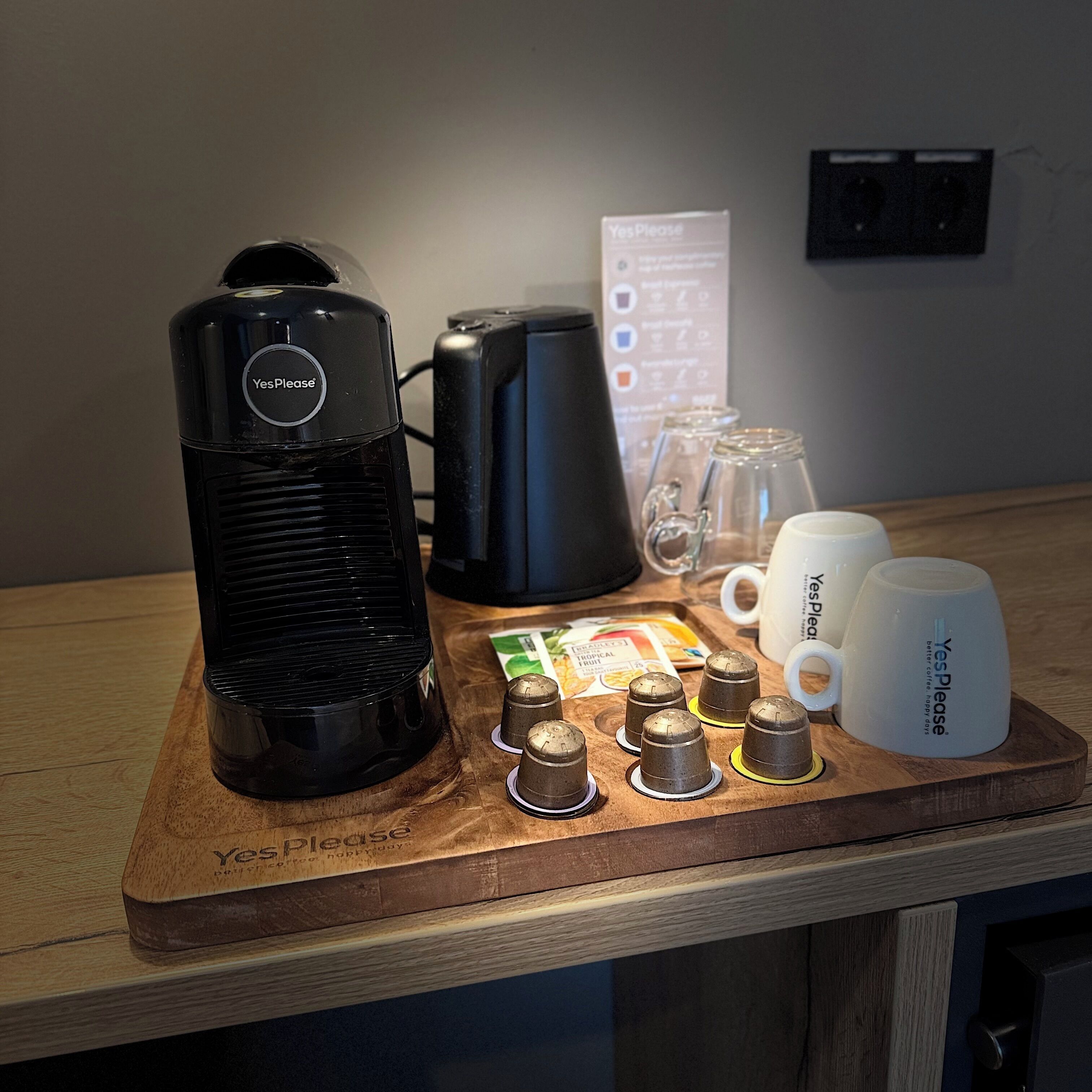 A coffee station with a Yes Please coffee maker, a black coffee pot, coffee cups, and coffee pods on a wooden tray.