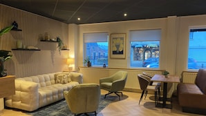 Een moderne woonkamer met een bank, fauteuils, een salontafel en uitzicht op de straat door grote ramen.