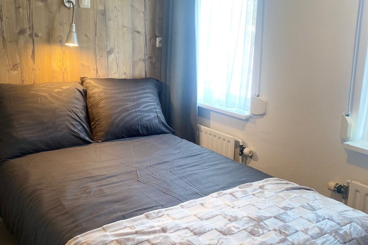 Ein Schlafzimmer mit einem hölzernen Kopfteil, einem Bett mit einer grauen und weißen Steudecke, einer Nachttischlampe und einem Fenster mit Vorhängen.