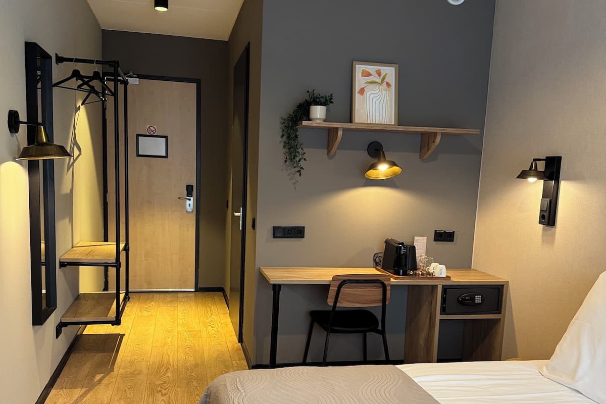 Ein modernes Hotelzimmer mit Bett, Schreibtisch und Stuhl. Eine kleine Ablagefläche zeigt eine Pflanze und ein Bild. Der Raum verfügt über einen Holzboden und eine Tür zu einem anderen Zimmer.