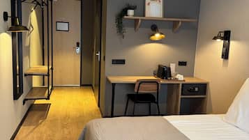 A modern hotel room with a bed, desk, and chair. There is a small shelf with a plant and a picture. The room has a wooden floor and a door leading to another room.