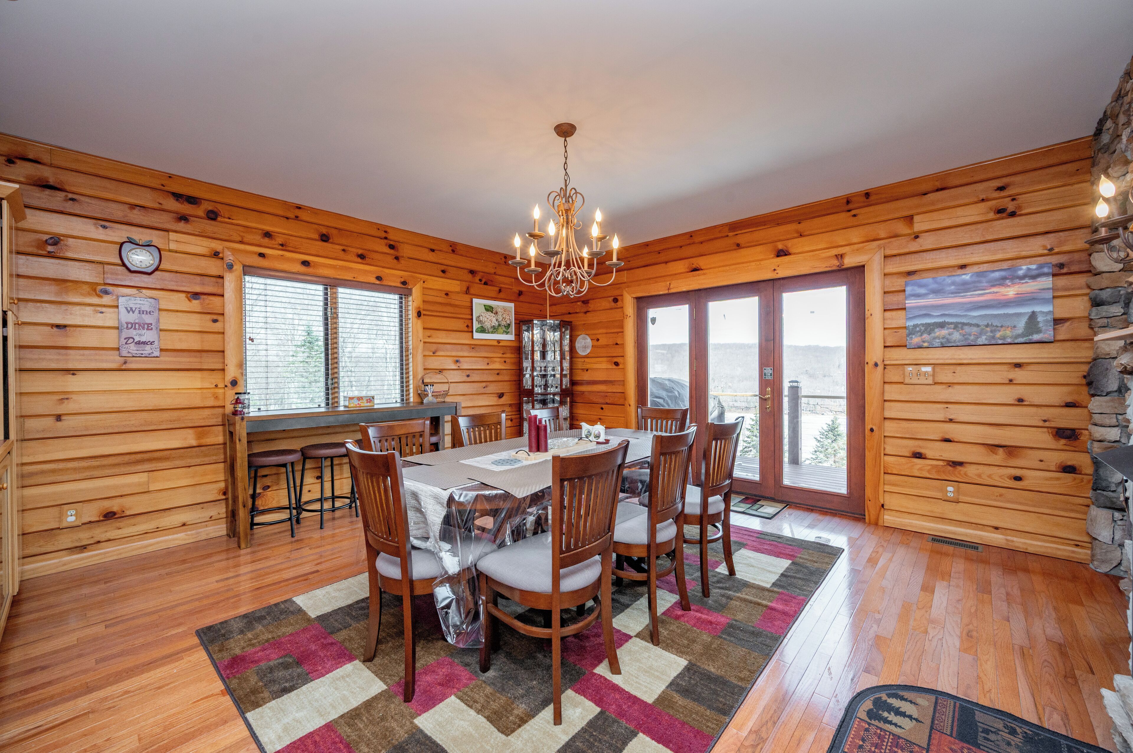 A wooden cabin with a dining area featuring a chandelier, a bar, and a large window.
