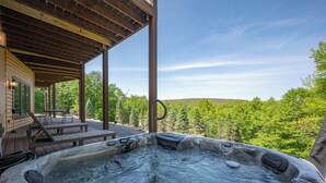 A hot tub with a wooden deck and outdoor furniture, surrounded by lush greenery and a clear sky.